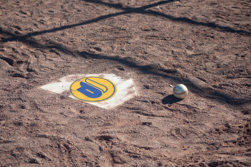 A baseball sits on gravel next to a home plate with a yellow and blue logo on it.