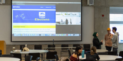 Image of a room with round tables and a screen. There are three people in the far right side of the room and two on the right side sitting down.