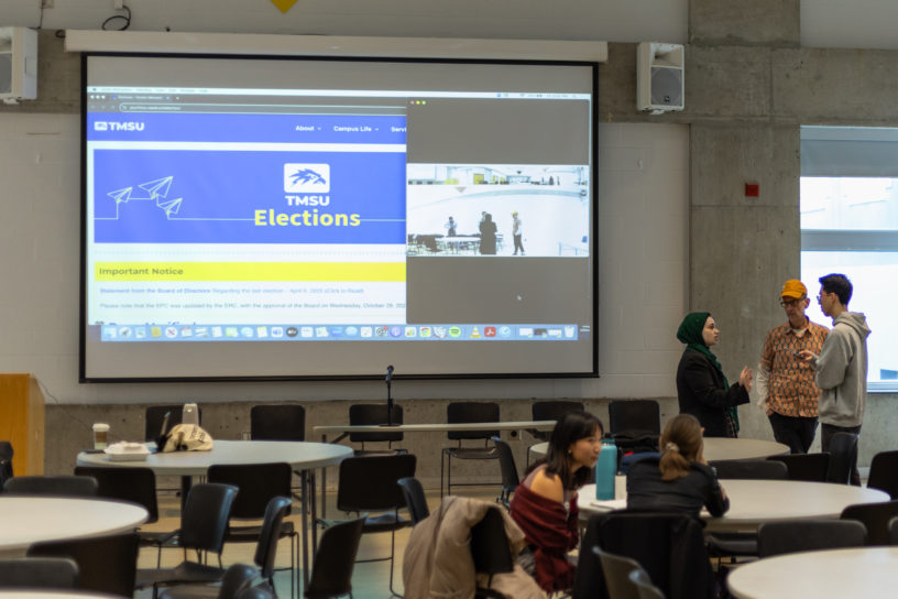 Image of a room with round tables and a screen. There are three people in the far right side of the room and two on the right side sitting down.