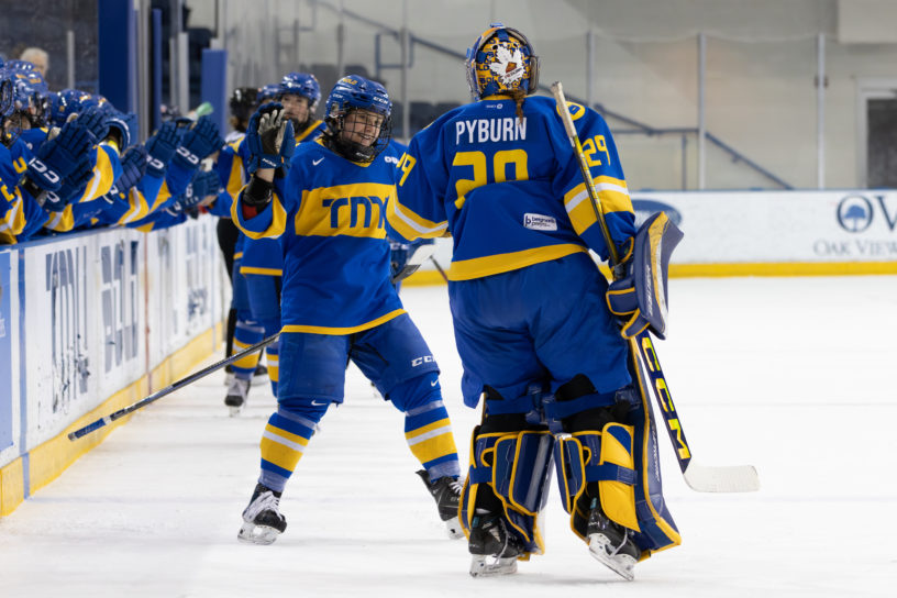 Two TMU Bold women's hockey players reaching for a high five in front on the TMU bench at the Mattamy Athletics Centre