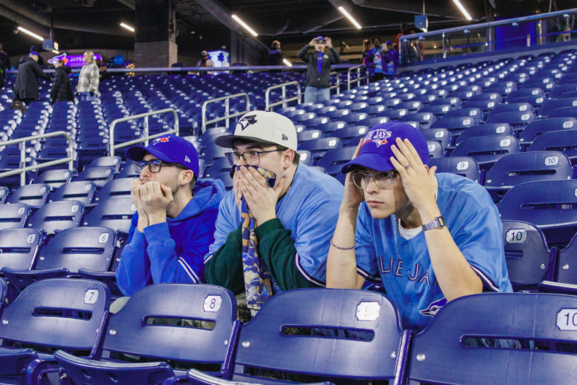 Three people sitting on blue seats in Blue Jays gear looking nervously forward.