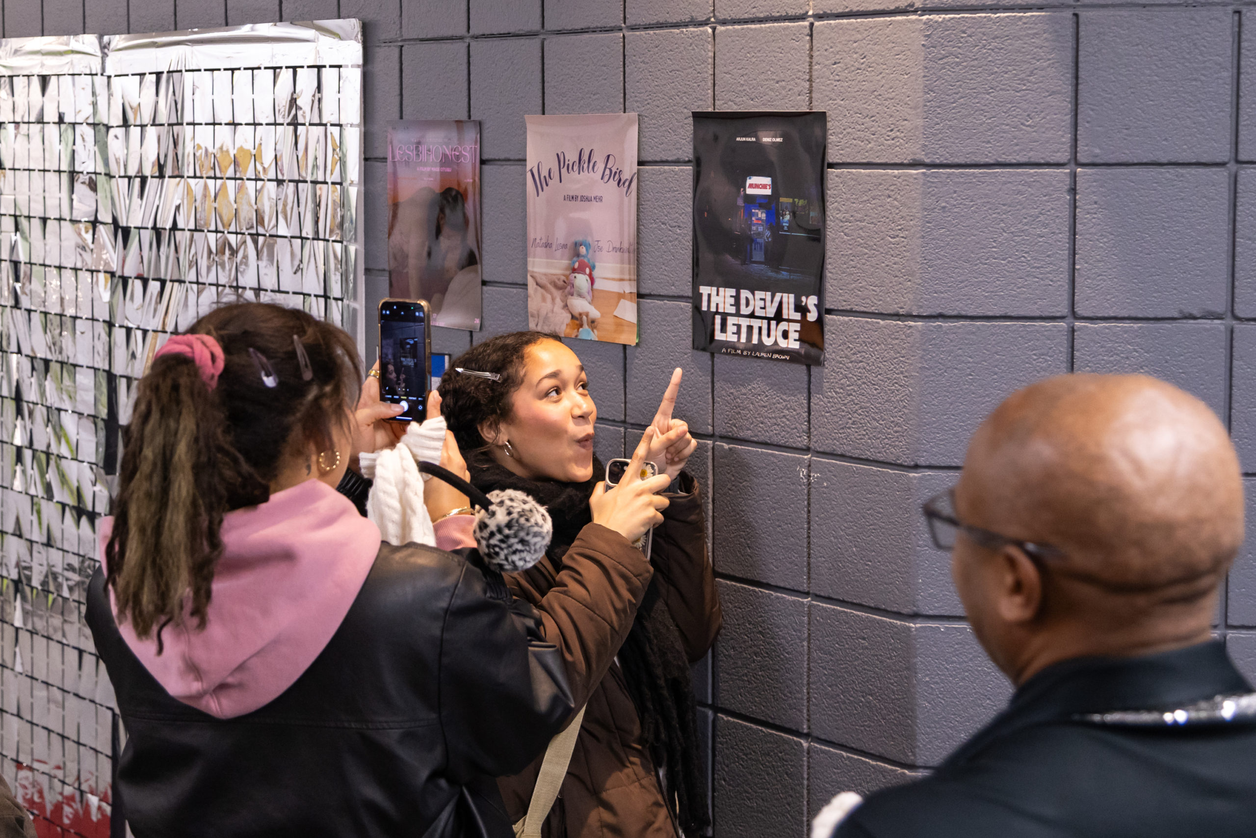 A person posing in front of a poster on a wall while another takes their photo on their iPhone.