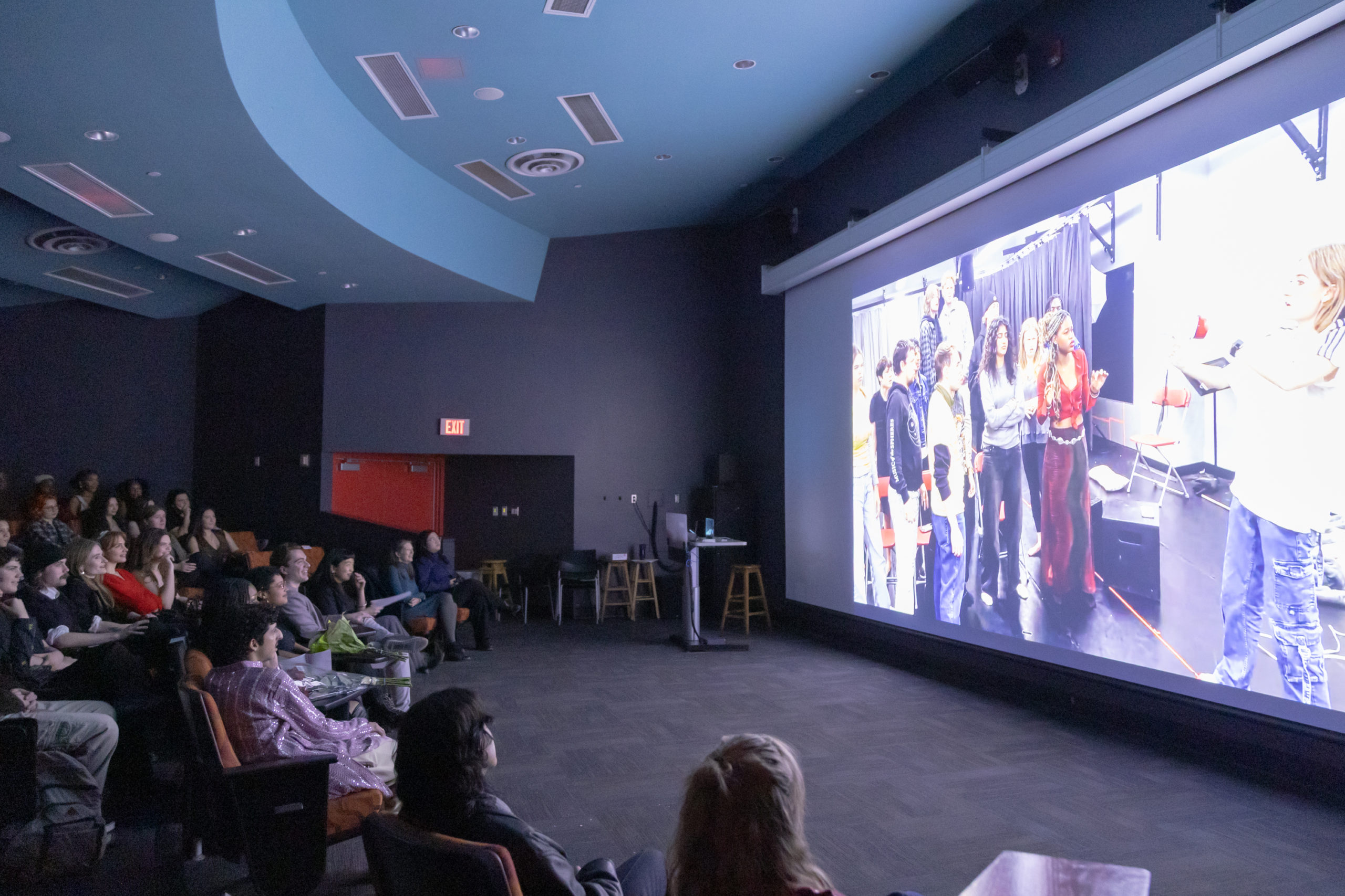 An audience watches a film on a large screen in a lecture room.