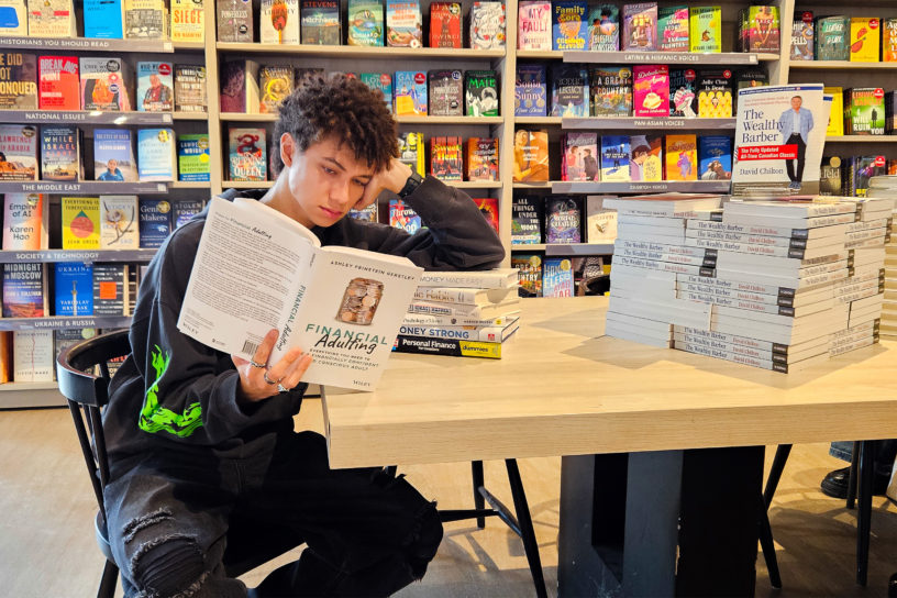 Image of a person reading a book titled "Financial Adulting" as they lean against a stack of books.