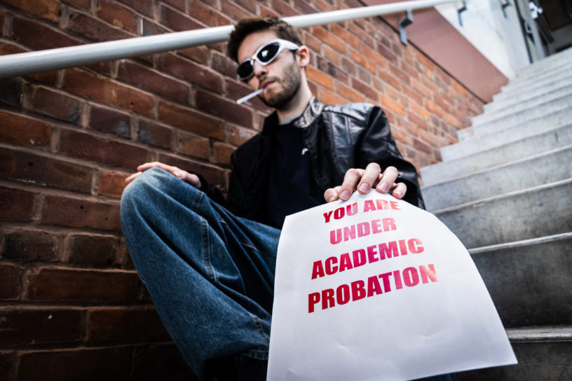 Photo of a man sitting on a staircase with a cigarette in his mouth, and holding a paper which reads "YOU ARE UNDER ACADEMIC PROBATION" in bold red text.
