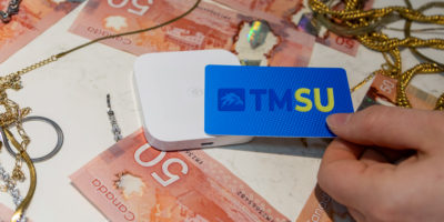 Image of someone tapping their card with a TMSU logo on it onto a card reader with $50 bills and gold chains around it.