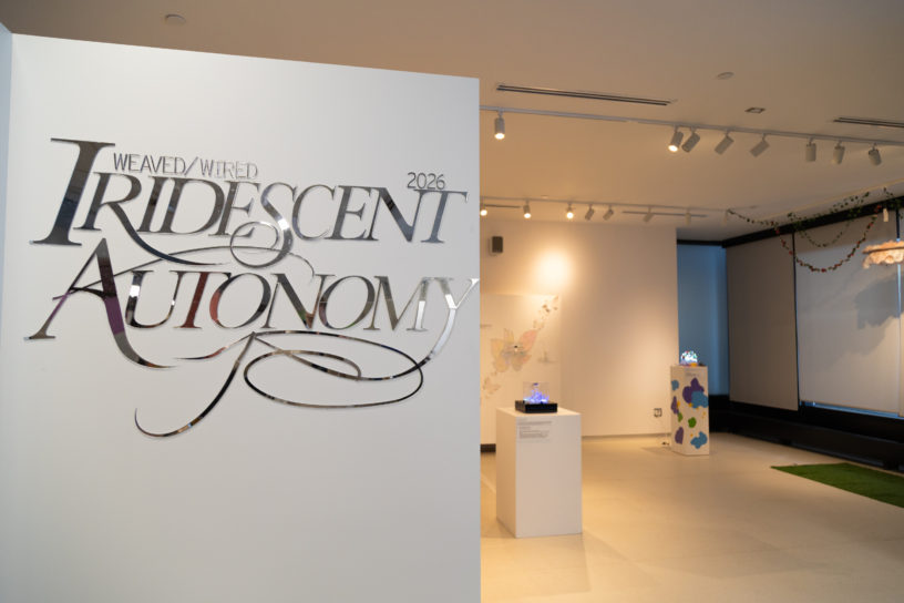 A sign that reads "Weaved/Wired Iridescent Autonomy" with the exhibit in the background
