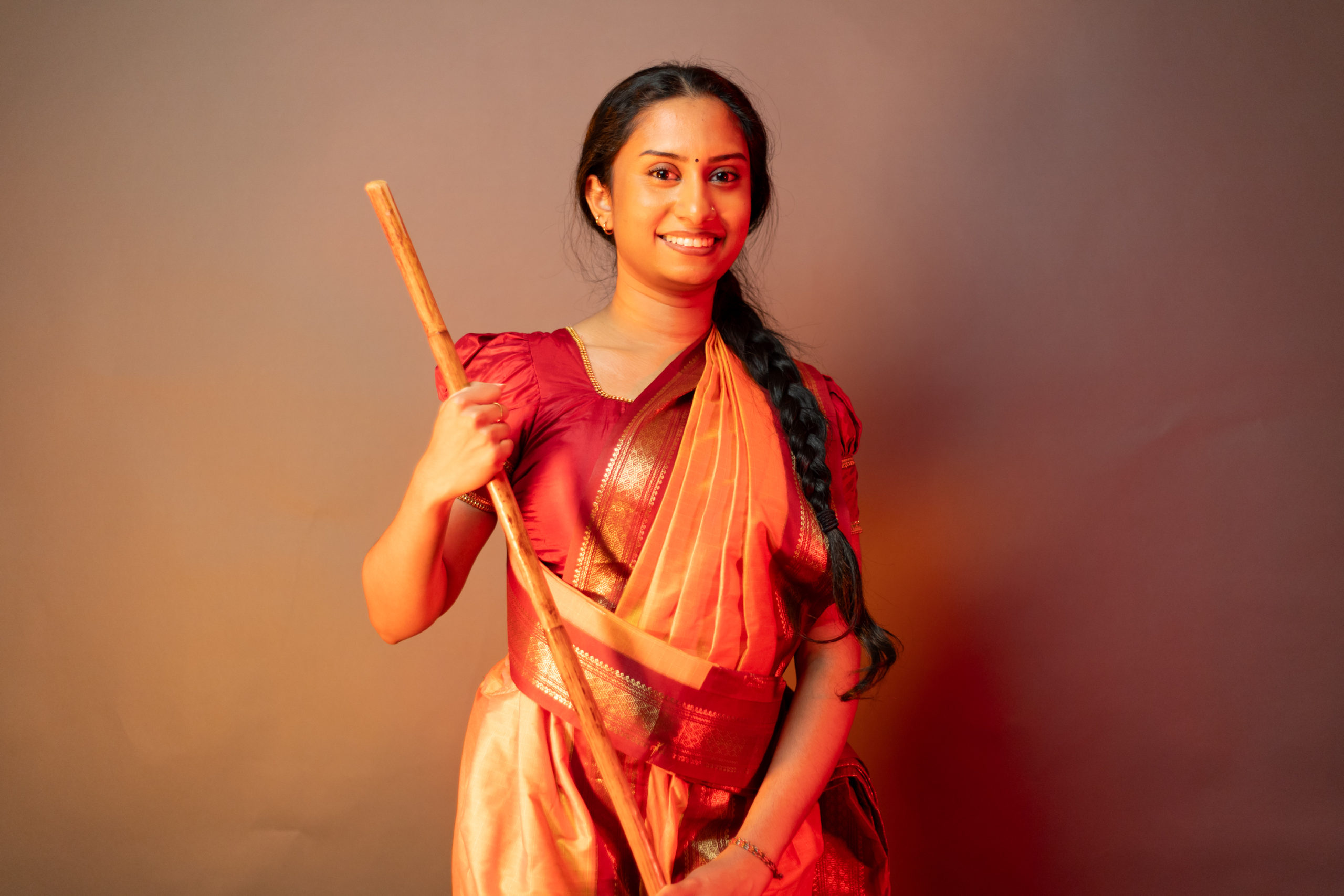 Aishvini Partheepan poses facing the camera holding her silambam staff across her body while wearing red and orange cultural clothing.