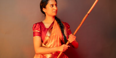 Aishvini Partheepan faces towards the right while holding up her silambam staff wearing red and orange cultural clothing.