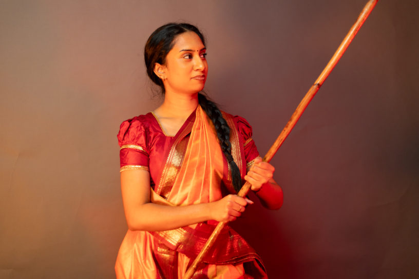 Aishvini Partheepan faces towards the right while holding up her silambam staff wearing red and orange cultural clothing.