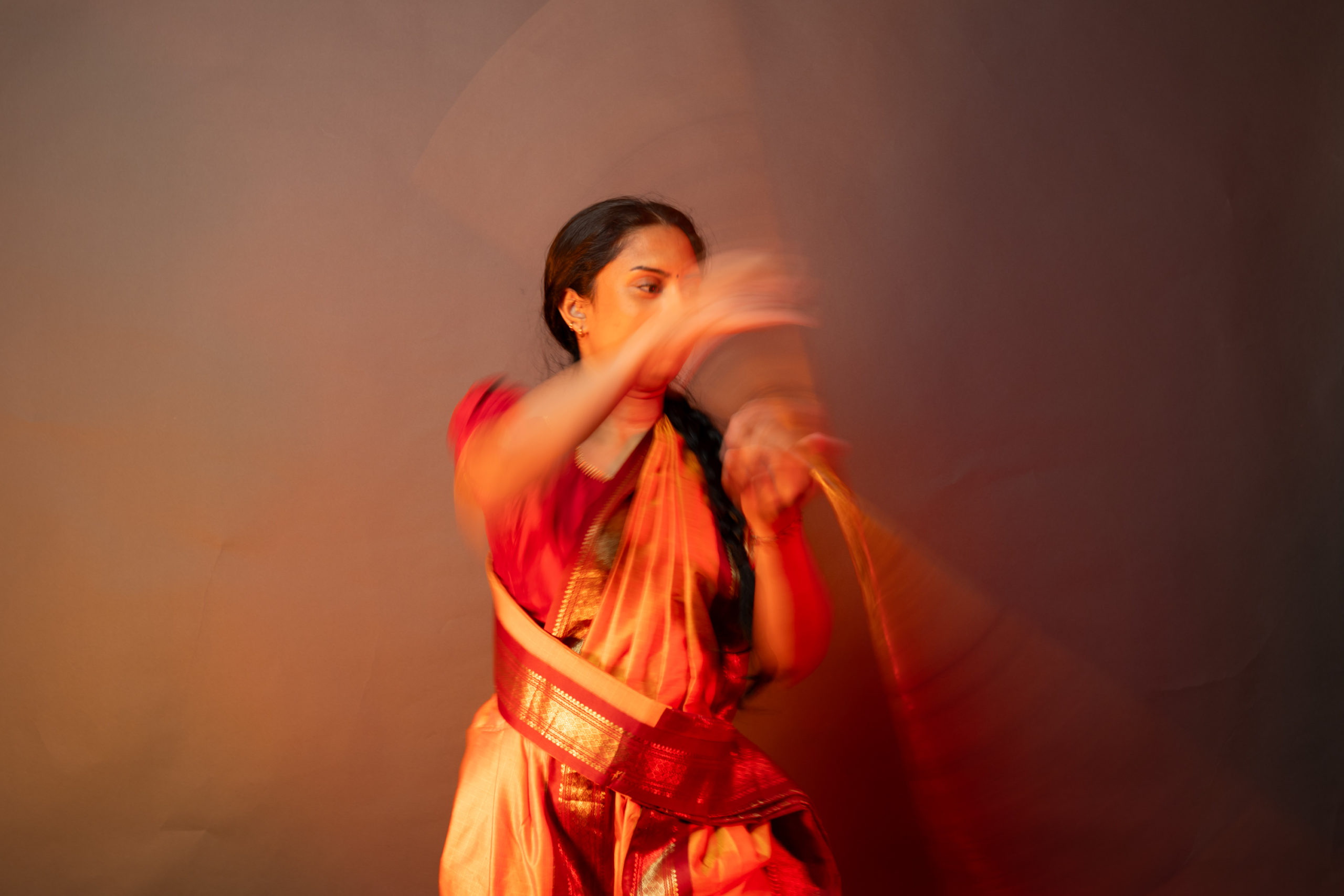 Aishvini Partheepan spins her silambam staff across her body while wearing red and orange cultural clothing.