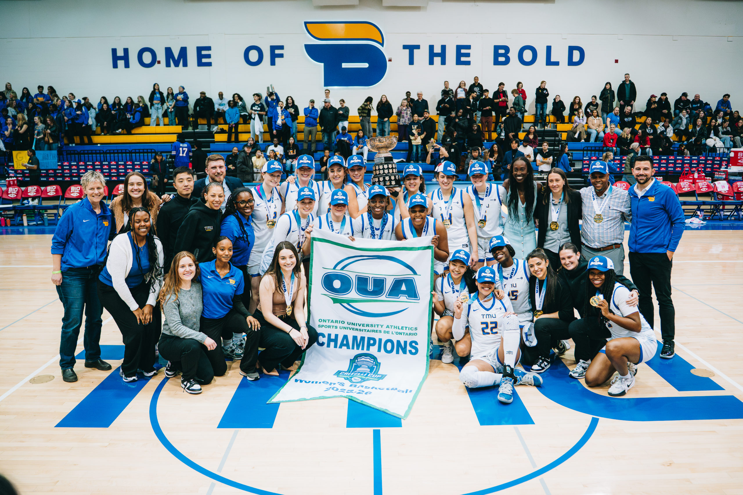 The TMU women's basketball team huddle together, hoisting the Critelli Cup and an OUA Championship banner