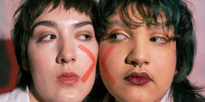 Two people with their faces pressed together with an X painted on their faces. They are looking at each other.