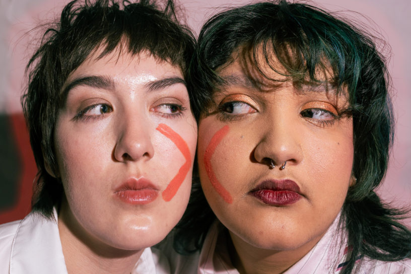 Two people with their faces pressed together with an X painted on their faces. They are looking at each other.