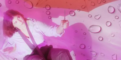 A person sits on a white backdrop holding a red umbrella. Rain drops are casted over the image.