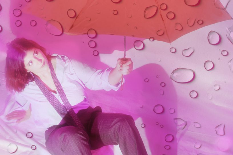 A person sits on a white backdrop holding a red umbrella. Rain drops are casted over the image.