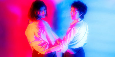 Two people stand with their hands in each other's back pockets. They are twisted towards the camera with red light coming from the left and blue from the right.
