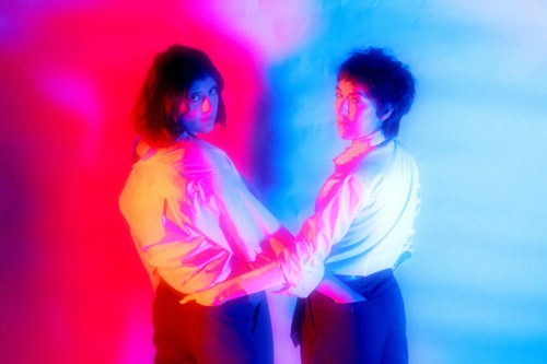 Two people stand with their hands in each other's back pockets. They are twisted towards the camera with red light coming from the left and blue from the right.