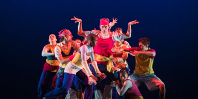 A group of dancers surround one person as they all move in different directions, they are in front of a blue background.