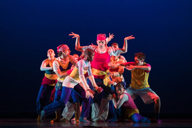 A group of dancers surround one person as they all move in different directions, they are in front of a blue background.