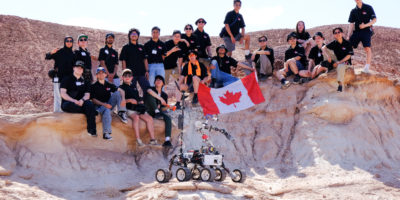 Image of a group of people posing for the camera outside in a rocky terrain with a robot.