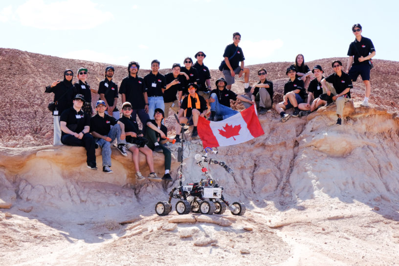 Image of a group of people posing for the camera outside in a rocky terrain with a robot.