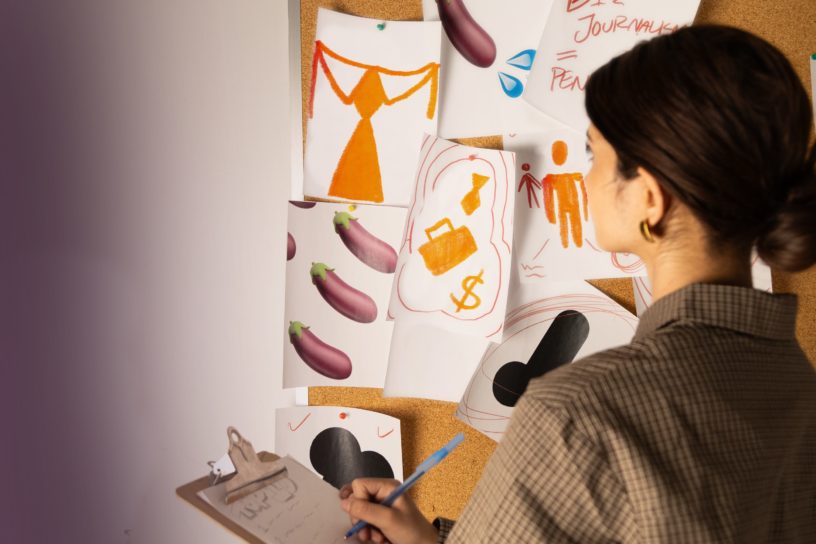 A woman holds a clipboard and studies a cork board full of images of eggplants, ties and a briefcase.