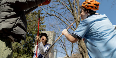 Three people set up for a jump ball with a basketball, a hockey stick and a broom while wearing a bike helmet and a baseball helmet.