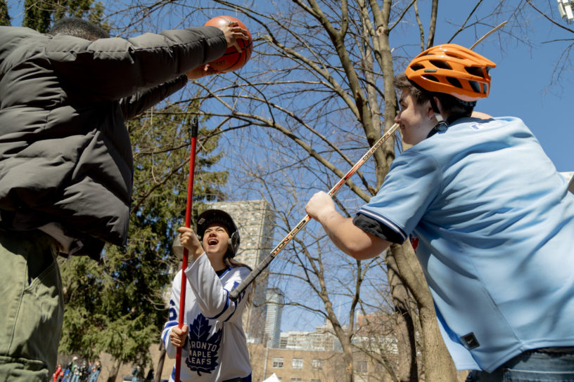Three people set up for a jump ball with a basketball, a hockey stick and a broom while wearing a bike helmet and a baseball helmet.