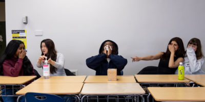 Students sitting at classroom desks with someone in the middle blowing their nose. The others on the right are laughing and using sanitizer while the two on the left are pointing and laughing.