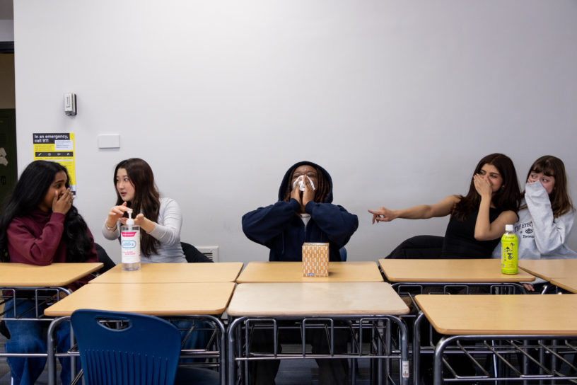 Students sitting at classroom desks with someone in the middle blowing their nose. The others on the right are laughing and using sanitizer while the two on the left are pointing and laughing.