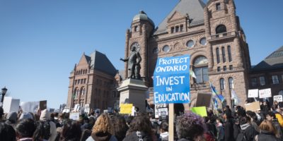 Protestors in front of the Ontario Legislative Building holding signs, one reads "PROTECT ONTARIO INVEST IN EDUCATION".