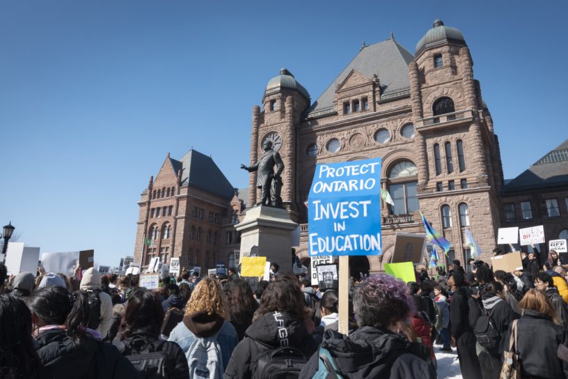Protestors in front of the Ontario Legislative Building holding signs, one reads "PROTECT ONTARIO INVEST IN EDUCATION".