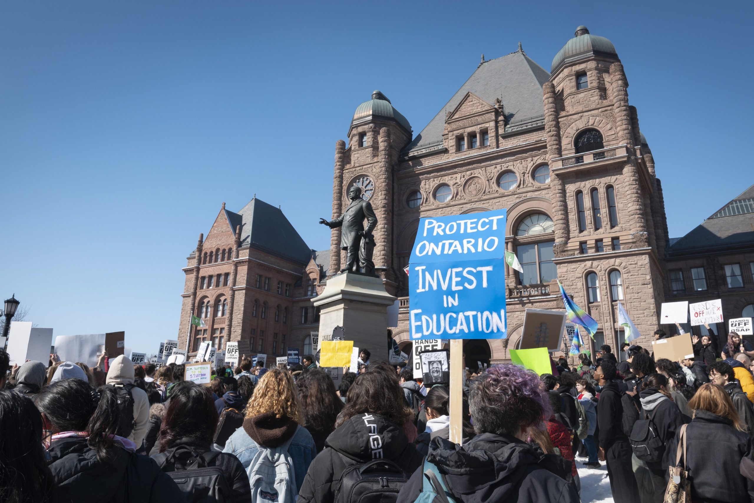 Protestors in front of the Ontario Legislative Building holding signs, one reads "PROTECT ONTARIO INVEST IN EDUCATION".