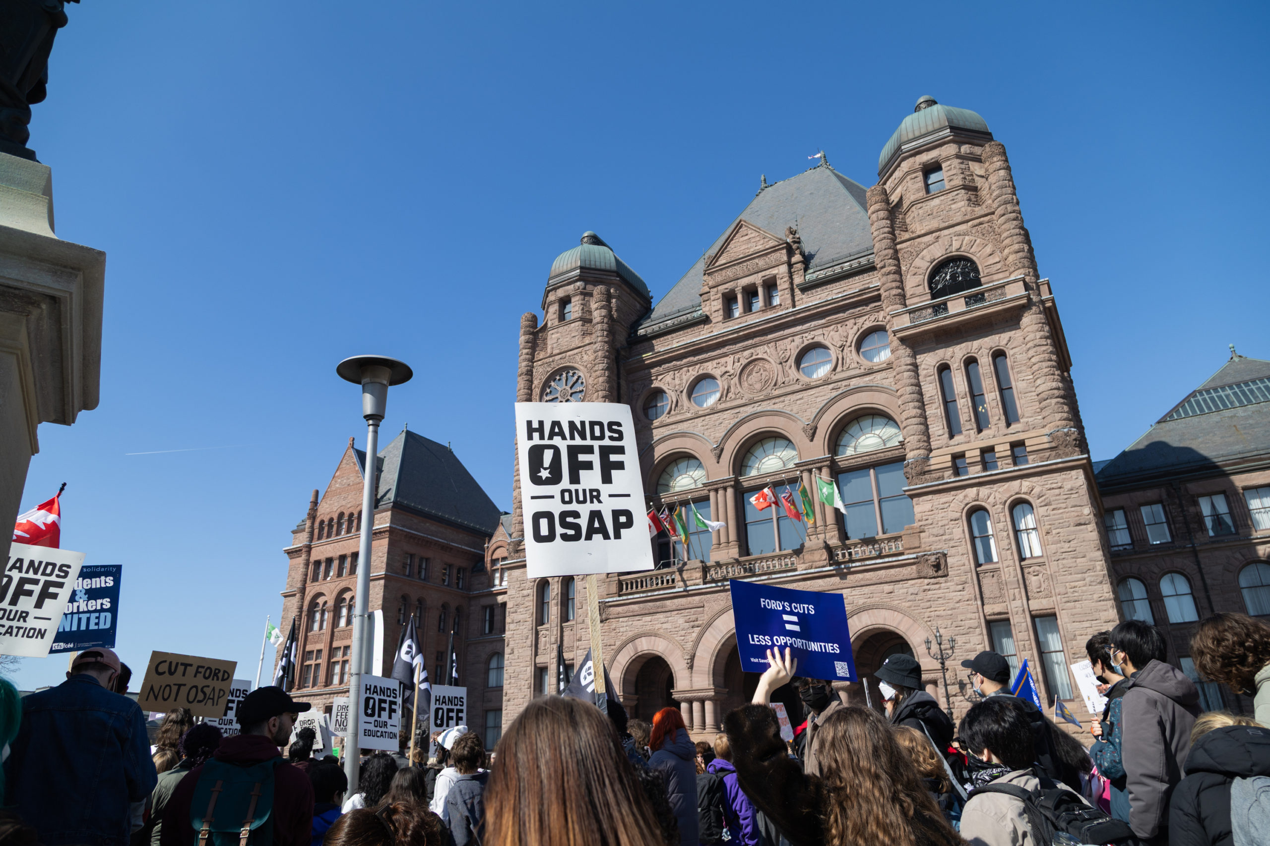 The back of people's heads who are holding signs in front of the Ontario Legislative Building.