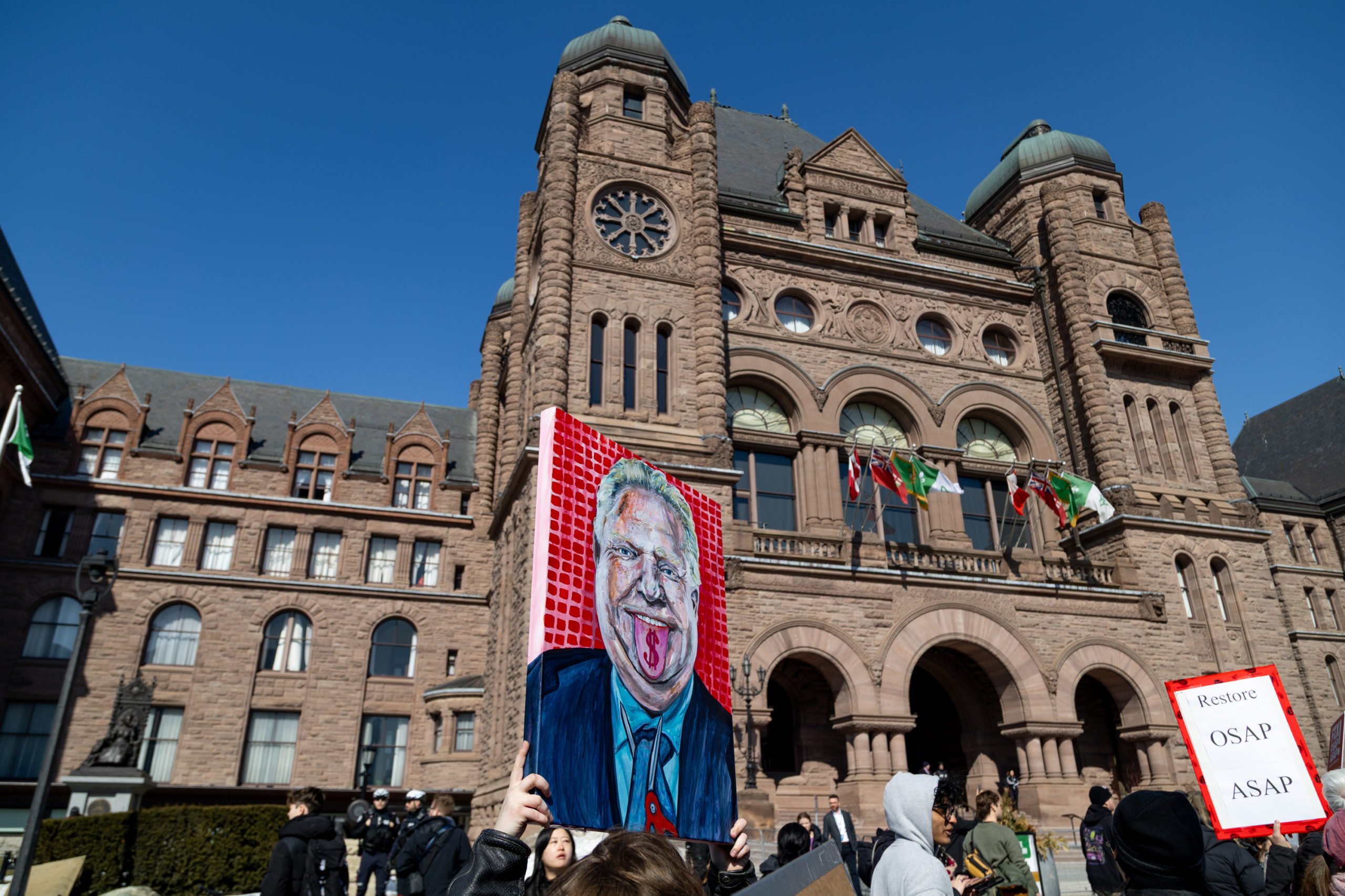Someone holding up a canvas with a painting of Doug Ford in front of the Ontario Legislative Building.