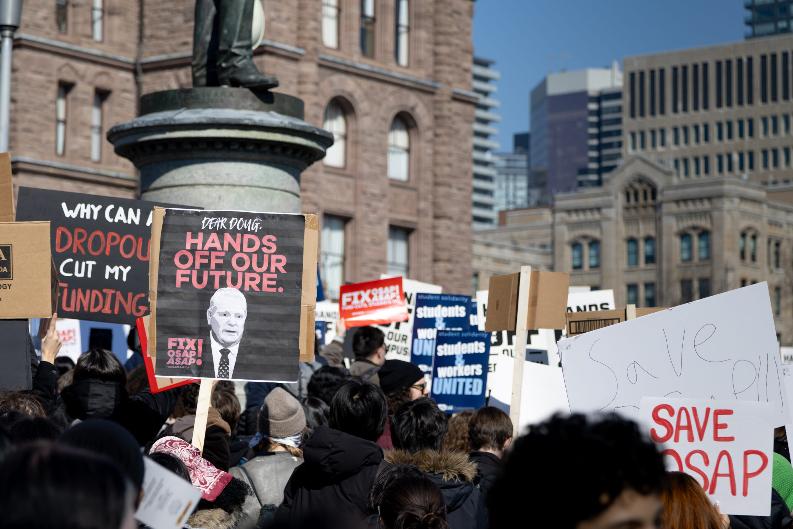 A group of people holding multiple signs in front of the Ontario Legislative Building.