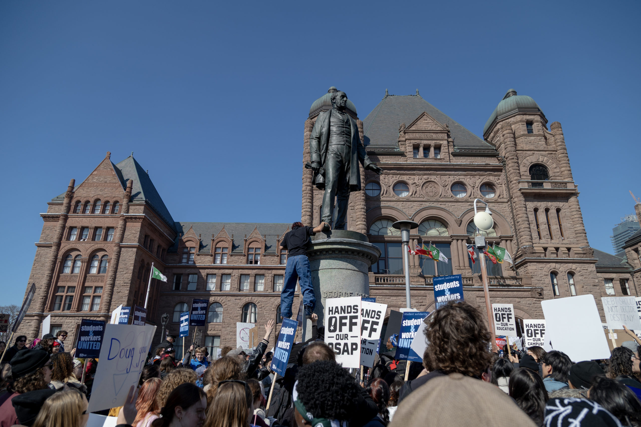 A group of people in front of the Ontario Legislative Building holding signs as someone climbs down from a statue.