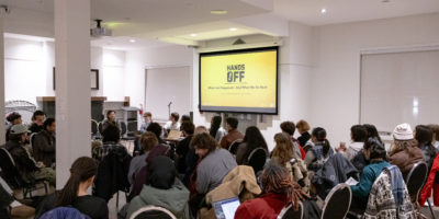A room filled with people sitting in chairs with a screen projecting the words "HANDS OFF".