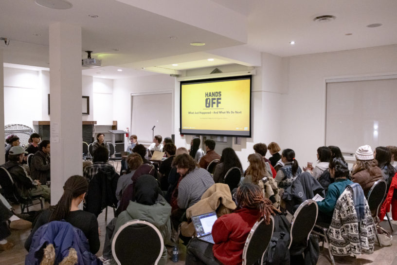A room filled with people sitting in chairs with a screen projecting the words "HANDS OFF".