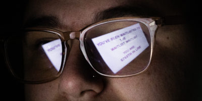 Photo of a man wearing glasses with the reflection of the glasses reading: "YOU'VE BEEN WAITLISTED TO THE WAITLIST WAITLIST" "YOU ARE: 6769TH IN LINE".