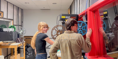 A man and women stand next to an old school film projector in the back room of a theatre.