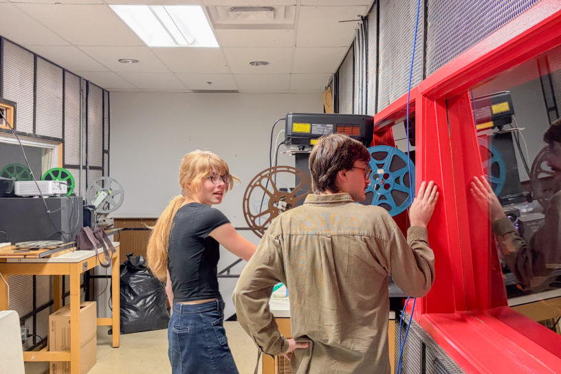 A man and women stand next to an old school film projector in the back room of a theatre.