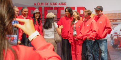 7 people in red shirts standing in front of a movie poster saying "Drifter a film by Sung Kang", with another person taking a photo of them
