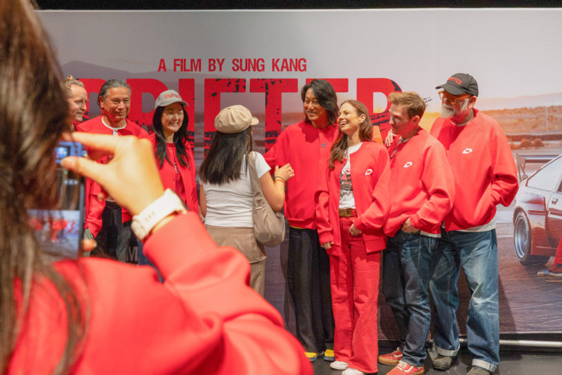 7 people in red shirts standing in front of a movie poster saying "Drifter a film by Sung Kang", with another person taking a photo of them