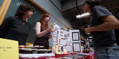 Picture of two students behind a table filled with cupcakes, a poster board and various items, speaking to someone on the other side of the table.