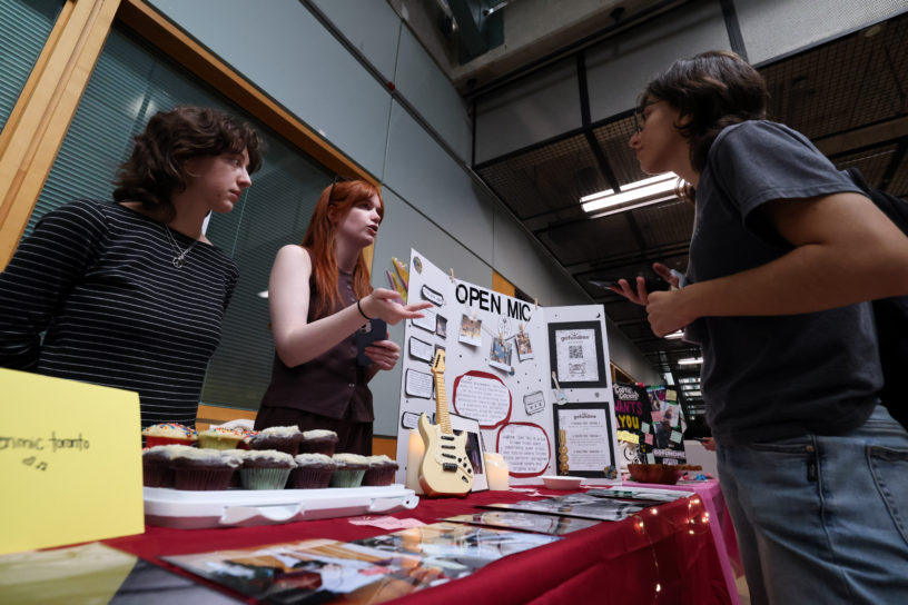 Picture of two students behind a table filled with cupcakes, a poster board and various items, speaking to someone on the other side of the table.