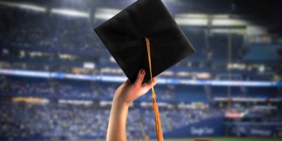 Photo of someone holding up a graduation cap on a background of a packed Rogers Centre.