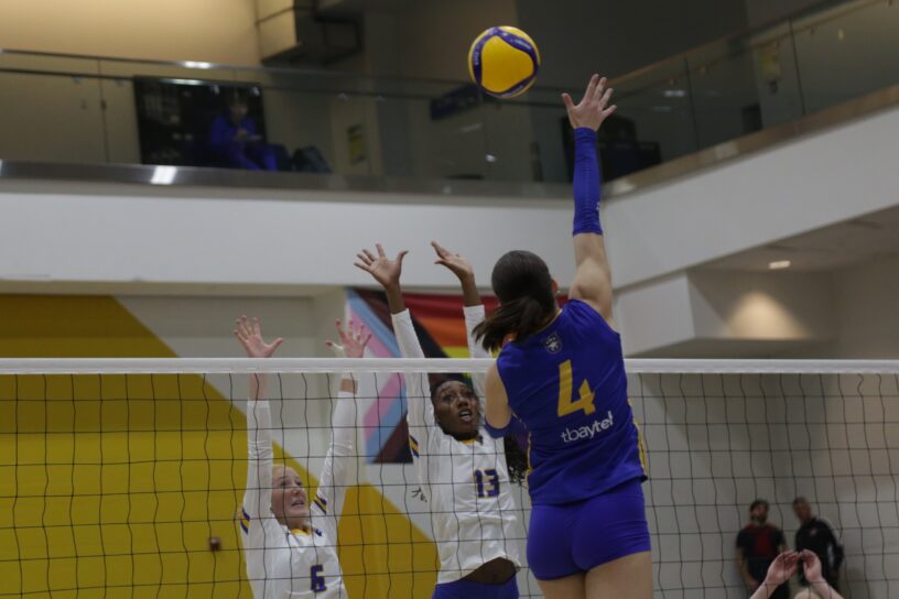 A Lakehead volleyball player attempts a kill in front of two TMU blockers