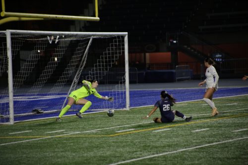 Bold goalkeeper dives sideways to save a ball in front of the net as a TMU player and U of T player approach